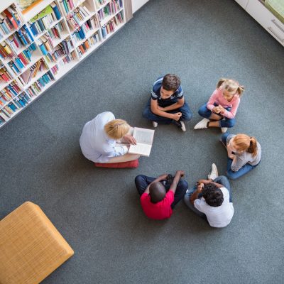 Teacher reading fairy tales to children sitting in a circle at library. Top view of librarian sitting with five multiethnic children on floor. Teacher reading book to cute girls and young boys at school.